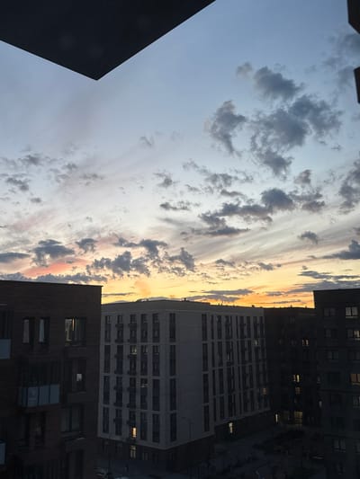 The image captures a city scene during sunset, with a dramatic sky filled with clouds. The sky transitions from a pale blue to warm hues of orange and yellow towards the horizon, illuminated by the setting sun. Scattered clouds, some dark and others wispy, streak across the sky, creating a dynamic visual.

Dominating the right side of the frame are several modern apartment buildings. The closest building in the foreground has a dark brick facade with numerous windows, some of which are lit, indicating occupants within. Behind it, a larger, lighter-colored building with a repeating pattern of balconies or windows stretches upwards. Further back, darker buildings are visible, with their windows also showing signs of interior lighting.

On the left side of the image, a large, dark, geometric structure, possibly another building or part of a construction, juts into the frame from the bottom left. Its sharp angles contrast with the softness of the sky.

The overall impression is of a late evening in an urban environment, where the natural beauty of the sunset is juxtaposed with the man-made structures of the city. The lighting suggests dusk or early evening, with the city lights beginning to compete with the fading daylight. The scene evokes a sense of calm and perhaps a touch of melancholy associated with the end of the day. No visible text is present in the image. The location is identified as Laikovo, Russia.