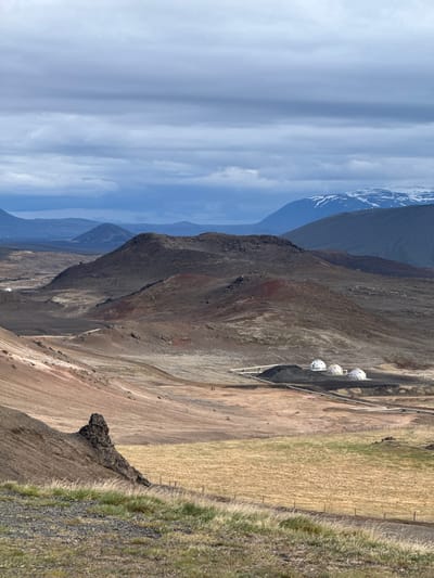 This image depicts a vast and rugged volcanic landscape in Reykjahlíð, Iceland, characterized by stark terrain and distant mountains under an overcast sky.

In the foreground, to the right, a section of dry, golden-brown grass transitions into a darker, rocky outcrop with sparse, yellowish-green vegetation. A faint fence line with thin posts can be discerned, separating different sections of the ground.

The midground features a broad, barren valley floor composed of light brown, reddish, and grey volcanic soil, showing signs of erosion and natural contours. Within this area, a notable cluster of three white, spherical or geodesic dome-like structures is visible, arranged in a row. These structures, possibly storage tanks or research facilities, are connected by subtle lines, likely pipes or pathways. Adjacent to them is a dark, rectangular patch, potentially a covered reservoir or foundation, with white supports or pipes running nearby.

Beyond the valley floor, the landscape ascends into several layers of mountains. The closest mountain range is a prominent dark reddish-brown and black, with a textured, rocky surface. Further in the background, successive mountain ranges appear in softer shades of blue and grey, with some of the most distant peaks distinctly capped with patches of snow.

The sky above is overcast, filled with layers of light blue and grey clouds that create diffused lighting across the scene. The overall impression is one of stark natural beauty and geological power. It is daytime, with soft, ambient light illuminating the scene. No human activity or visible text is present.