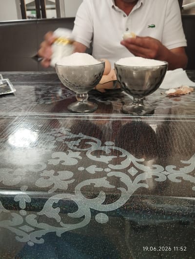 In Sasyk-Bulak, Kyrgyzstan, on June 19, 2026, at 11:52, an individual is shown in a close-up shot, with a focus on a table setting. The person, wearing a white polo shirt with a green crocodile logo, is in the process of peeling a boiled egg, holding a piece of it in their right hand. In front of the individual are two glass dessert bowls filled with what appears to be granular sugar or salt. The bowls are placed on a dark, reflective table with a decorative pattern. To the right of the bowls, there is a pile of discarded eggshells and a white napkin. The background shows a dark, cushioned seating area and a blurred view of what might be a room interior. The overall scene suggests a meal or snack preparation.