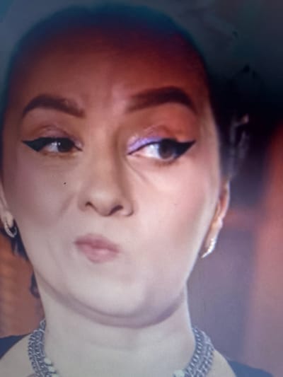 A close-up shot of a person's face. The person has dark eyes with winged eyeliner, pronounced eyebrows, and pink lips. Their skin tone appears to be light brown. Part of a silver chain is visible around the neck. The photo is rotated, with the face appearing tilted towards the right. It is not possible to determine the location or any further details based on the image.