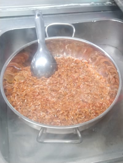 A stainless steel bowl sits within a stainless steel sink. The bowl is filled with small, orange shrimp, with a stainless steel ladle resting in the shrimp. The scene is located within Chu Văn An Ward, Vietnam.