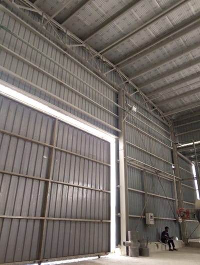 The image shows a large metal door, partially open, leading into a large warehouse-like structure. The walls and ceiling of the warehouse are made of corrugated metal. The door is made of multiple sections, and the walls have rows of corrugated metal panels. The ceiling has an insulated layer of white material.  A man in dark clothes is sitting on the floor near a white wall, facing a piece of equipment on his right.  A white cable is attached to the ceiling, running down the wall and to the piece of equipment, with a loose end on the floor.  The coordinates place the scene in a large industrial or commercial building in Pangasia, Bangladesh.