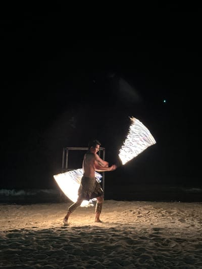 The image shows a shirtless male standing on a sandy beach, presumably at night in Hat Khai Muk, Thailand. He is performing a fire dance with two triangular torches. The beach is dark, but the sand is illuminated by the fire. In the background, there is a dark structure. A small, bright point of light is visible in the upper left. The image is rotated 90 degrees clockwise.