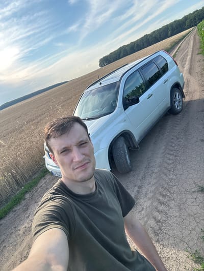 A man takes a selfie on a dirt road in Slobidka-Zalisetska, Ukraine. The man is wearing a green t-shirt and is standing in front of a white SUV. The SUV is parked on the side of the road, which is lined with a field of wheat on the left and a field of corn on the right. In the background, a forest can be seen. The sky is blue with some white clouds. The license plate of the car is partially visible and reads "EX 8662".