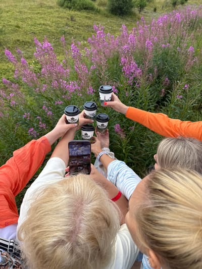 In an outdoor setting with a background of green grass and purple flowering plants, several individuals are gathered. One person is holding a smartphone, taking a photograph or video of the scene. Several hands are extended, each holding a small, black-lidded coffee cup with white branding. The cups appear to be for takeaway beverages. The attire suggests a casual gathering, with visible clothing including an orange shirt and a blue and white striped shirt. The overall impression is a moment captured during an outing in a natural environment. The specific location of T/B Seminskii Pereval, Russia, is not discernible from the visual elements of the image itself.