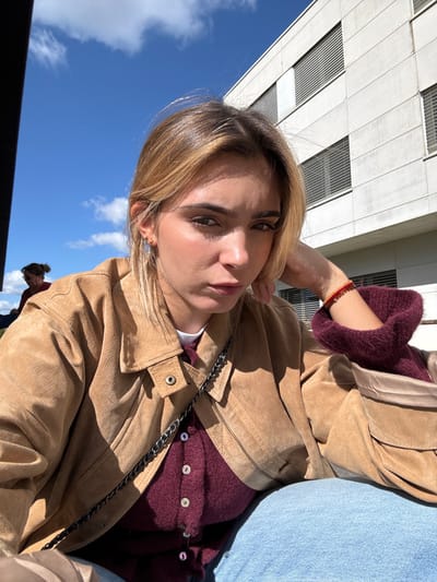 The image displays a young female subject seated outdoors in Saint-Priest, France, under a clear blue sky with scattered white clouds. She is positioned centrally, looking directly at the camera with a neutral expression, her right hand supporting her jawline. Her hair is light brown with blonde highlights. She wears a tan suede-like jacket over a burgundy knit cardigan with white buttons, and a white undershirt, paired with light blue denim jeans. A black chain-strap bag is visible across her torso. Small gold hoop earrings are present, and multiple red and gold bracelets adorn her left wrist. In the soft-focus background, a second individual with dark hair tied back and wearing a red top is partially visible to the left. A large, multi-story modern building with a light-colored facade featuring horizontal window or ventilation panels occupies the right background. The scene is illuminated by strong direct sunlight, casting distinct shadows.