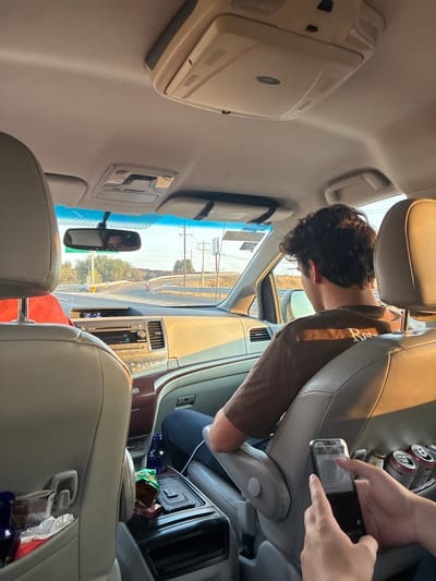 The image provides an interior view of a multi-passenger vehicle, likely a minivan or SUV, in motion during daylight hours in Santa Ana Xalmimilulco, Mexico. Warm, direct sunlight illuminates the cabin from the right.

In the immediate foreground, two hands, belonging to a rear passenger, operate a dark-cased smartphone. To the right, a seat-back storage area holds at least three metallic beverage cans.

The back of a front passenger is visible, characterized by dark, curly hair. This individual wears a brown short-sleeved t-shirt with light text and dark blue pants. Their right arm rests on the armrest, and a white charging cable extends from their lap towards the center console. The front passenger seat is upholstered in a light, possibly beige, material.

The center console, situated between the front seats, contains a blue plastic bottle, a crumpled green snack bag, and a red packet. Behind the driver's seat, a mesh pocket contains a blue plastic bottle and white paper items.

The vehicle's dashboard features a beige upper section, a metallic-silver center stack with a radio/CD player, and dark wood trim. An overhead console, equipped with a retracted screen, is mounted on the ceiling. The rearview mirror reflects a partial view of the driver's face, showing dark hair.

Through the windshield, an asphalt road curves to the left. A light-colored scooter or small motorcycle with a rider is visible on this curve. Beyond the roadway, utility poles with overhead lines are visible against a clear sky, with trees and foliage in the distant background.