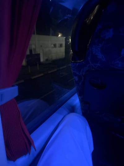 The image displays a nighttime scene observed from inside a vehicle, located in Starovoitove, Ukraine. The interior of the vehicle is illuminated by a strong blue ambient light. On the left, a pleated red curtain is drawn back by a light-colored plastic or fabric tie. In the lower center, the legs of an individual, clad in white fabric, are visible, prominently illuminated by the blue light. To the right, a dark blue patterned seatback, featuring a light-colored ornamental design, is partially visible. Through the window, a dark exterior environment is present. A multi-story, light-colored building with three visible square windows is dimly illuminated in the mid-ground. In front of the building, a paved surface is visible, exhibiting horizontal light-colored markings and a dark, pole-mounted object or sign. No individuals are distinctly visible outside the vehicle, and no overt actions or interactions are depicted.