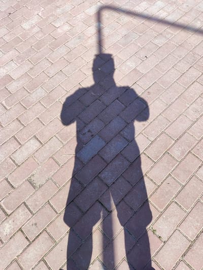 A person's shadow is cast on a brick-paved walkway. The shadow depicts a person from the neck down, with arms crossed over their chest. The walkway is made of reddish-brown rectangular bricks laid in a herringbone pattern. The bricks appear slightly weathered and have some small patches of green growth in the grout lines. The shadow is elongated, suggesting sunlight is coming from a low angle. A faint shadow of an overhead structure, possibly a bar or fence, is also visible extending from the top of the person's head. The environment appears to be outdoors, likely in a public space given the paving. The location is Snezhinsk, Russia.