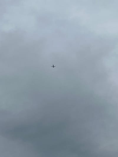 A small, dark airplane is visible in the center of the frame against a grey, overcast sky. The plane appears to be a propeller-driven aircraft, with its wings extended horizontally and a tail fin. It is captured in flight, with its silhouette clearly defined against the diffused light of the clouds. The sky is uniformly cloudy, suggesting a dull or possibly rainy day. There are no discernible landmarks or environmental details that would suggest a specific location beyond the general context of being outdoors under a cloudy sky. The image appears to have been taken from below, looking up at the airplane. The overall mood is neutral, with the focus solely on the solitary aircraft in the vast expanse of the sky. There is no visible text in the image.