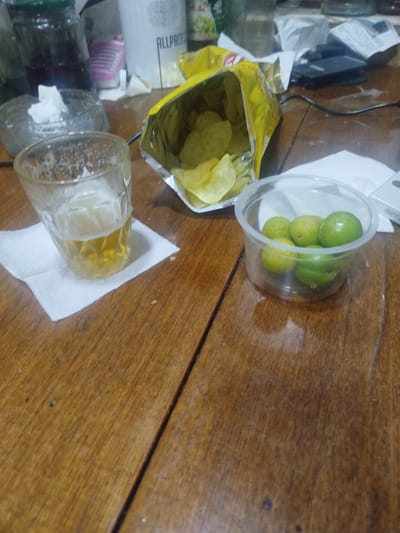 The image depicts a close-up, slightly blurry view of a wooden table with various items. In the foreground, to the left, is a glass containing a yellow liquid, likely beer, resting on a white paper napkin. To the right of the glass, an open bag of potato chips is spilling out its contents. Further to the right, a clear plastic container holds several small, green fruits, possibly limes or small lemons. In the background, out of focus, are other objects including a large dark bottle, a white container with text, and what appears to be electronic equipment. The lighting suggests an indoor setting, likely in the evening or a dimly lit room. There are no people visible in the image. The scene appears to be set in a casual environment, possibly a home or a small gathering. There is no information present in the image that would indicate its location in Sabunchu, Azerbaijan.
