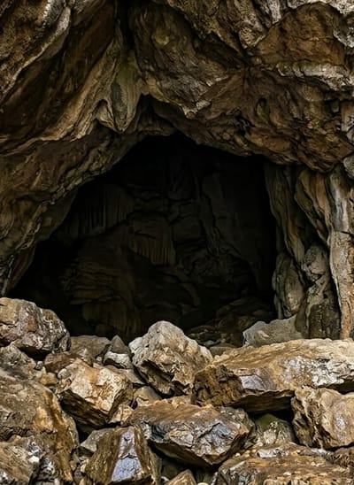 The image depicts the entrance of a cave, characterized by a rugged, natural rock formation. The foreground is dominated by a dense cluster of large, irregularly shaped rocks, some appearing wet and reflective, suggesting recent moisture. These rocks vary in color from light brown to dark earth tones, with visible textures and natural imperfections.
Beyond the rocky entrance, the cave opens into a dark, cavernous interior. The walls and ceiling of the cave exhibit complex geological formations, including stalactites hanging from above and layered rock structures. The lighting within the cave is dim, creating deep shadows that obscure finer details of the inner space.
The scene is set in the natural environment of Penungkiren, Indonesia, known for its geological features. There are no people or animals visible in the image. The overall impression is one of a raw, natural cave entrance, untouched by human intervention.