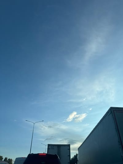 The image captures a clear blue sky with scattered wispy clouds. In the foreground, the tops of several large trucks are visible, suggesting they are in transit. One truck on the right side has a prominent red stripe and partial text, possibly a brand name. In the middle ground, a street lamp with two lights stands tall. On the left, another street lamp is partially visible. A sliver of the moon can be seen in the upper left quadrant of the sky. The overall scene suggests a daytime view, possibly from a highway or a congested road. The image does not contain any people or any identifiable landmarks that would pinpoint the location to Krutitsy, Russia.
