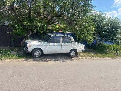 A white, older model sedan is parked on a dirt and grass shoulder next to an asphalt road. The car is positioned under the shade of a large, leafy tree with a wooden fence to its left and a blue fence to its right. The vehicle has a roof rack and appears to be slightly weathered with some rust visible. In the background, houses with tiled roofs are visible, as well as power lines stretching across a bright blue sky with scattered clouds. The environment suggests a residential area in Pereiaslavske, Ukraine, on a clear, sunny day. No people are visible in the image.