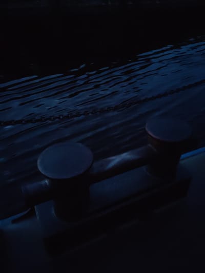 The image depicts a dark, moody scene with a focus on a boat cleat on a dock. The water is dark and rippled, reflecting the dim light, with a chain visible partially submerged. The cleat itself is a dark, metallic object with two rounded bollards mounted on a rectangular base. It appears to be made of heavy-duty material. The overall impression is one of a quiet, possibly nighttime, maritime setting. The location is identified as Annenskii Most, Russia. There are no people or animals present in the image.