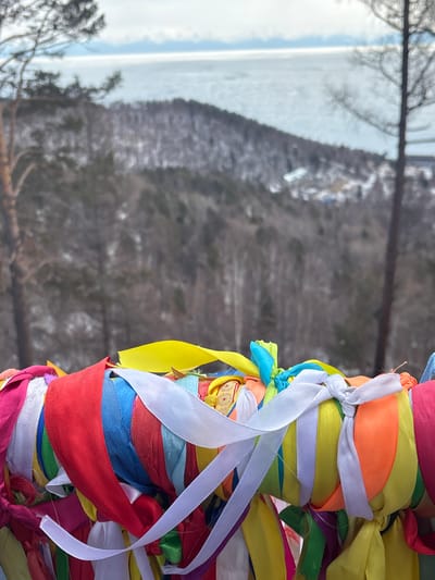 The image displays a dense foreground of numerous multi-colored fabric ribbons tied to an unseen horizontal structure, likely a railing. The ribbons vary in width and material, featuring prominent colors such as red, yellow, blue, green, orange, pink, white, and light blue, secured with various knots and bows. The background, significantly blurred, depicts a winter or early spring landscape in Nikola, Russia. A snow-dusted, forested hillside with mostly bare or sparsely leaved trees descends towards a large body of water, identified as Lake Baikal, which appears partially frozen or covered in ice. Small, indistinct structures are visible on the distant part of the hillside. The sky above the lake is overcast. Out-of-focus portions of tree trunks are visible along the left and right edges of the frame. No individuals are present in the scene.
