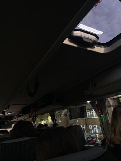 The image shows the interior of a bus. The bus is parked on a street in Potters Bar, United Kingdom. The coordinates provided place the bus near a residential area. There are several people on the bus, most of them are seated. The windows of the bus show views of houses and a street. The bus has a sunroof open and a monitor on the back of the seat in front. The roof of the bus has a dark grey interior.  A bright light from outside shines through the sunroof.  There is a handle on the ceiling of the bus.  The image is dark and appears to have been taken at night or early in the morning.