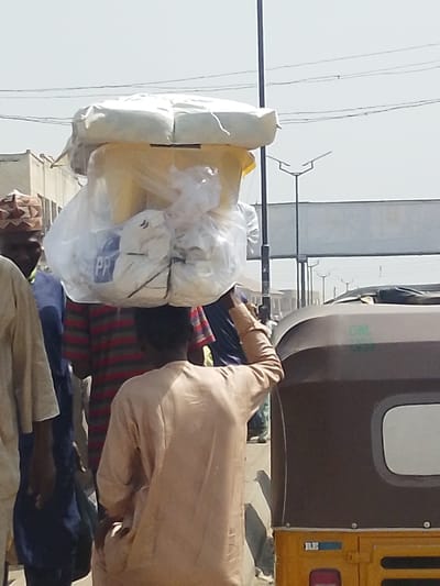 The image depicts a bright, sunlit outdoor scene in Fagge D1, Nigeria, showing a street or market environment. In the foreground, a person, viewed from behind, wears a light tan long-sleeved garment and carries a substantial, multi-component load on their head. The load consists of a large, light-colored bundled item atop a yellow container, further wrapped and supplemented with clear plastic bags containing white and light-colored materials, one bag showing the letters "PP". The individual's right arm is raised, with the hand near the load, possibly steadying it.
To the immediate left of the central figure, a man with dark skin is partially visible, wearing a brown patterned cap and a blue over-garment. Further left, another person wears a red and dark-striped shirt. To the right, the rear upper section of a brown and yellow three-wheeled vehicle, consistent with an auto-rickshaw, is visible, showing a small window and a green logo on its brown canopy.
The background features a clear, pale sky with overhead power lines. A tall, dark pole stands vertically near the center. A series of modern streetlights with distinctive upward-angled luminaires extends into the distance along what appears to be a road. Light-colored buildings are visible in the far left background, and a large, rectangular structure, possibly a billboard, is present in the distant background. The ground surface is indistinct but suggests a paved or dirt road.