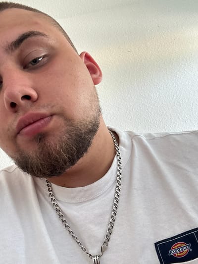 The image is a close-up, low-angle, eye-level shot of a young adult male. He has short hair, a stubble beard, and is wearing a white t-shirt with a Dickies logo on the chest. A silver chain necklace is visible around his neck. The background is a plain white textured wall. The image was taken in Schkopau, Germany.