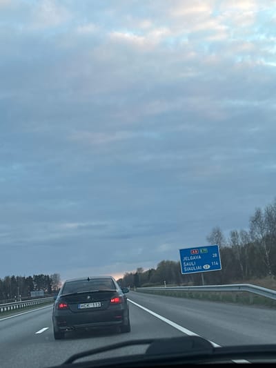 The image captures a perspective from inside a vehicle traveling on a highway, looking forward at a black BMW sedan directly ahead in the right lane. The car, possibly a 5-series model, displays the license plate "HCK 113".
To the left of the highway, a blue road sign is prominently visible. It indicates route numbers "A8" and "E77" at the top. Below these, destinations are listed with their distances: "JELGAVA" is 28 units away, and "ŠIAULIAI" is 114 units away, accompanied by a "LT" symbol with an arrow, signifying the direction towards Lithuania. The highway features a white dashed lane marking and a metal guardrail along its side. Beyond the guardrail, a sparse forest with bare trees lines the road.
The sky is overcast with soft, diffused light, suggesting either late afternoon or early morning hours. The overall setting is an open road in a semi-rural environment, consistent with the location context of Jaunolaine, Latvia, indicating travel on a main route towards the Lithuanian border.