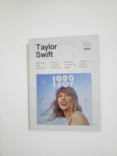 The scene is located in Al Haouafate, Morocco. An upright, rectangular print is affixed to or leaning against a plain, off-white wall. The print itself features a two-tone design. The upper section has a white background with numerous small, dark speckles, resembling a distressed texture. It displays the name "Taylor Swift" in large, dark gray sans-serif font at the top-left. Below this, song titles are listed in three columns: "Blank Space," "Style," "Shake It Off" in the first; "Anti-Hero," "Love Story," "All Too Well" in the second; and "Enchanted," "The Great War," "august" (lowercase) in the third. In the top-right corner, a stylized globe icon appears above the numbers "1989," with "Ready For It?", "Gorgeous," and "Bad Blood" listed below "1989" in a fourth column.

The lower section of the print presents a light blue, textured background, reminiscent of a sky or a faded photograph, also with dark speckles. Large white numbers "1989" are prominent in the center, with "TAYLOR'S VERSION" in smaller white text directly beneath. The primary subject in this section is a fair-skinned female figure, identified as Taylor Swift, positioned in the lower-right quadrant, smiling broadly with visible teeth, and looking slightly upwards and to her left. Her shoulder-length, wavy, light brown hair appears wind-blown. She is wearing a light-colored, possibly white, strapless or thin-strapped garment. Several white seagulls are depicted in various stages of flight across the blue background, primarily above and to the left of the female figure. The entire print appears to be flat against the wall, with no significant damage or creases visible.