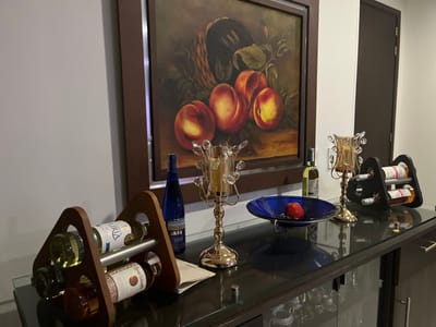 This image displays an interior scene, likely within a private residence or commercial establishment in Floridablanca, Colombia, though a specific street address or public landmark is not identifiable from the visual data. No individuals or actions are depicted within the frame.
The central focus is a dark-colored console table or buffet with a reflective glass surface, positioned against a plain, light-colored wall. Above the table, a large, dark-framed still-life painting features a composition of peaches or similar stone fruit, possibly within a basket, against a dark background.
Arranged on the glass tabletop are various objects: On the far left, a triangular wooden wine rack secures four wine bottles horizontally, with visible labels including "Aliria" and "Gran Valiosa" on two bottles. Next to this, a tall, slender dark blue bottle stands upright. Centrally positioned are two ornate, gold-colored metallic candle holders, each adorned with clear, crystal-like elements, flanking a dark blue, irregular-rimmed glass bowl. This bowl contains one red apple and a small cluster of dark berries. To the right, an upright clear glass bottle of white wine is visible next to a dark-colored, triangular wine rack holding two additional wine bottles horizontally. A dark brown door is partially visible on the far right side of the frame, indicating an adjacent room or area. The scene is illuminated by interior lighting.