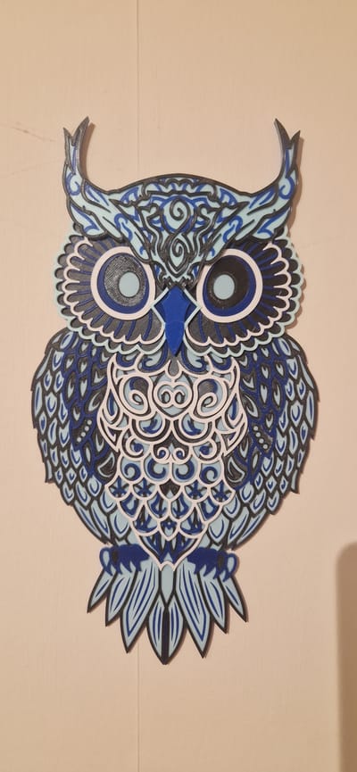 A layered owl decoration in blue, black, and white is affixed to a pale, textured wall. The owl is depicted front-facing, with large circular eyes, stylized feathers, and pointed ear tufts. The decoration appears to be made of a rigid material, possibly wood or plastic, with intricate cut-out details. The wall has a slight texture and is illuminated from the side, casting a subtle shadow to the right of the owl. The overall impression is of a decorative piece mounted indoors. No specific location in the city of South Holland, United Kingdom, can be determined from the image.