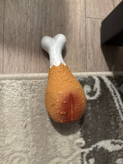 A dog toy shaped like a chicken leg rests on a wood-grain floor and a patterned rug. The toy has a white, bone-shaped end and an orange, textured body. A dark, rectangular object is partially visible in the upper right corner of the image. The scene does not provide any information about the city of Perm, Russia.