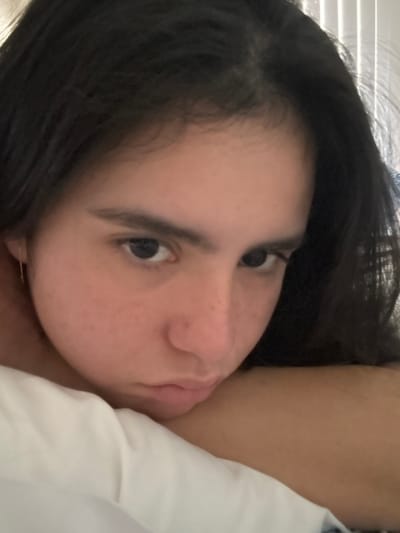 A medium close-up, slightly low-angle photograph features a young female subject. She has dark, shoulder-length hair, dark brown eyes, and a medium complexion with faint freckling across the nose and upper cheeks. A thin, gold-colored threader earring is visible in her left ear. Her head rests on a light brown surface, likely an arm, with a section of white fabric, consistent with a pillow or sheet, partially obscuring her lower left face and shoulder. Her gaze is directed towards the camera, and her mouth is closed in a neutral or slightly pensive expression. The background displays vertical white window blinds, indicating an indoor setting with ambient light. The scene is located in Santiago, Mexico.