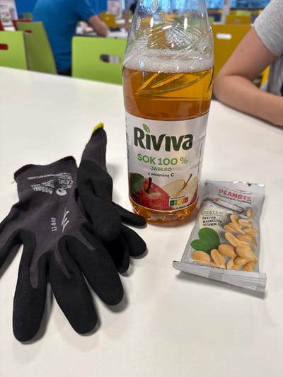 The image depicts a close-up, eye-level view of a white table. Positioned on the table are a pair of black work gloves, a bottle of Riviva apple juice with the label partially visible, and a small packet of peanuts. The gloves are placed to the left of the image, with their fingers curled. The bottle of juice is upright to the right of the gloves, and the peanut packet is in front of the juice bottle. In the blurred background, two individuals are seated at tables, with one wearing a blue shirt and the other a grey shirt. The environment appears to be an indoor common area, possibly a break room or cafeteria, with brightly colored chairs visible in the background. There is no information in the image to suggest the location is in Chociule, Poland.