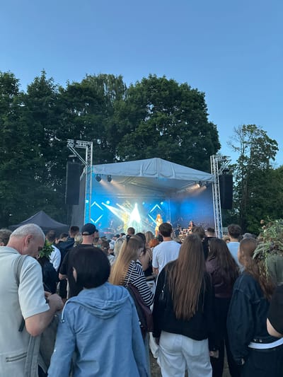 This media file depicts an outdoor concert scene with a crowd of people gathered to watch a band perform on a stage. The stage is equipped with lights and speakers, and the band members are visible playing their instruments. The event appears to be taking place in the evening, as indicated by the twilight sky. The surrounding area is filled with trees, suggesting a park or outdoor venue. The mood seems lively and engaged, with the audience focused on the performance.