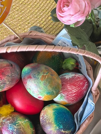 The image depicts a close-up view of an Easter basket filled with brightly colored Easter eggs. The eggs are dyed in various shades of red, green, blue, and yellow, some with a speckled or crackled pattern. The basket is lined with a blue-and-white patterned cloth. A pink artificial flower and green leaves are attached to the basket's exterior. A pale pink handle curves into the frame. Yellow and green decorative chicks are also present. The basket sits on a woven surface, likely straw. There is no evidence to suggest it is located in the city of Bretton, United Kingdom.