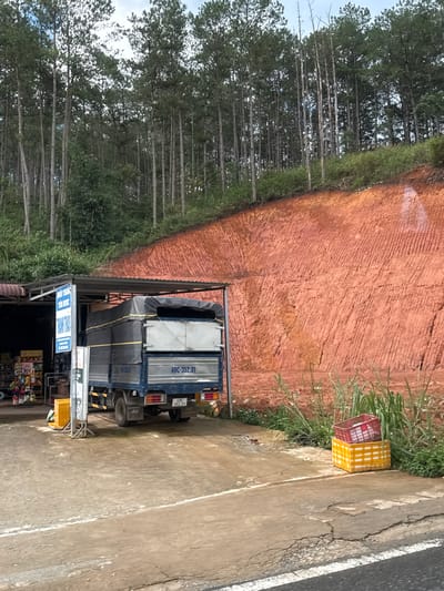 The image depicts a roadside scene in Klong Klanh, Vietnam, characterized by a truck parked next to a roadside stall, with a large, reddish-brown dirt embankment behind it. The embankment is marked with parallel striations, suggesting it is a cut or prepared surface. A dense pine forest covers the top of the embankment, with the trees extending into the background and reaching towards a cloudy sky.
In the foreground, a blue truck with a tarp-covered cargo bed is parked under a corrugated metal awning. The truck displays a license plate that reads "49C-352.31" and below it "49C 352.31". To the left of the truck, a roadside stall is visible, with shelves stocked with various products, including bottles and packaged goods. A yellow container is positioned near the stall.
To the right of the truck, near the edge of the road, a stack of two plastic crates is placed on the ground. The bottom crate is yellow and white, and the top crate is red. Tall green grass and smaller white wildflowers grow in patches along the embankment and the roadside. The road surface in the foreground is cracked and worn, with a white painted line running along its edge. The overall impression is of a functional, working roadside environment in a somewhat rural or semi-rural setting.