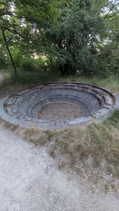 The image shows a circular, tiered concrete structure in a wooded area. The structure is approximately circular with multiple concentric levels descending towards a central floor covered in gravel and dirt. The outer edge of the structure is a concrete rim with some chipped and broken sections. The tiers are constructed from grey bricks that show signs of wear and weathering. Surrounding the structure is dry grass and vegetation, with trees visible in the background. A gravel path leads away from the structure to the left. The lighting suggests it is either early morning or late afternoon. The scene appears to be an outdoor public space, possibly a historical or recreational site. There are no people or animals visible in the image.