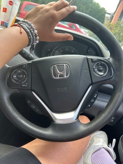 The image shows the interior of a car, specifically the driver's side. The steering wheel is prominently displayed with the Honda logo in the center. On the steering wheel, there are various control buttons. A hand is holding the steering wheel and a straw from a McDonald's Coca-Cola drink can be seen. The arm is adorned with multiple bracelets. A leg and a shoe with a purple sock are visible on the right.