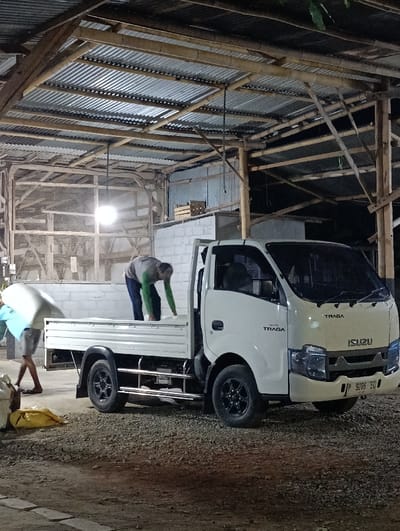 The image shows a white Isuzu truck parked under a corrugated metal roof structure supported by wooden beams in Kecamatan Kapongan, Indonesia (latitude: -7.673, longitude: 114.061).  A man in a green shirt is loading something into the truck bed.  Another man, wearing a blue shirt, is walking away from the truck.  The truck has a "TRAGA" logo on the side and the license plate number "9268 EQ". The ground surrounding the truck is covered in gravel.  The image appears to have been taken in a rural area, potentially a construction site or a loading dock.  The scene suggests the loading of materials for a construction project.