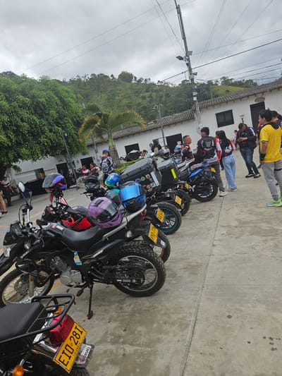 This image depicts an outdoor scene in Bucarasica, Colombia, featuring a gathering of people and numerous motorcycles under an overcast sky. The foreground and midground are dominated by parked motorcycles, predominantly black, with various colored helmets (purple, blue, pink/black) resting on their seats or handlebars. Several motorcycles display license plates, including "EIO 281 COLOMBIA," "KXZ 66N," and "YYQ 77A." Some motorcycles are identifiable as Suzuki models, with "DR 150" visible on one. Many bikes are equipped with top cases and luggage racks.

Approximately 15-20 individuals are scattered throughout the midground, standing in groups and conversing. Most are adults in casual attire, some wearing jackets with motorcycle club or brand logos, such as "DRAGON ROJOS" and "SUZUKI." A person in a yellow shirt and gray pants stands prominently on the far right, while another in a black Adidas jacket focuses on a mobile phone. A woman in light blue jeans and a dark top stands near a motorcycle further back.

In the background, a row of white, single-story buildings with dark, possibly tiled, roofs and dark windows/doors is visible. One building displays a sign that reads "JUEGOS POOL." Lush green foliage, including large trees and a palm tree, flanks the buildings and extends to a prominent, tree-covered hill that forms the distant backdrop. A tall utility pole with multiple wires, light fixtures, and attached boxes stands centrally in the midground, extending upwards against the gray sky. The ground surface is a light gray concrete or paved area, exhibiting minor stains and cracks.