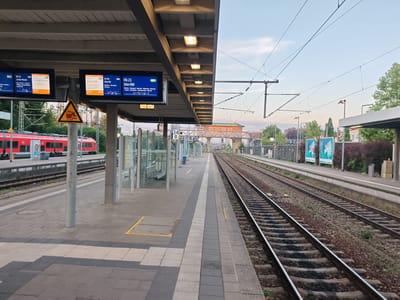 The image depicts a view of a train station platform in Memmingen, Germany, under a clear sky with a gentle sunset glow. On the left, a red train is parked at a platform, with a digital display above showing train schedules. The platform is paved with grey tiles and marked with yellow lines. A yellow triangular warning sign with a silhouette of a person falling is visible on a pole. On the right, multiple train tracks run parallel to the platform, disappearing into the distance. The tracks are lined with gravel and ballast. Overhead, a complex network of electrical wires for the trains is suspended from poles. In the background, there are buildings, trees, and advertising posters. The overall scene suggests a quiet moment at the train station as evening approaches.