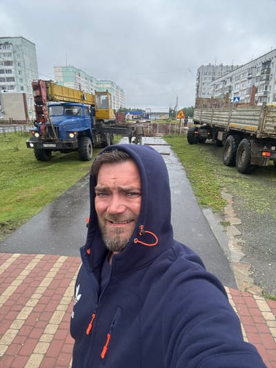 A man wearing a dark blue hooded sweatshirt with orange drawstrings is taking a selfie in Usinsk, Russia. He has a beard and is smiling. Behind him, a blue crane truck is parked on a grassy area next to a paved walkway with red and beige pavers. Further in the background, a flatbed truck is also parked. Multiple multi-story apartment buildings are visible on either side of the street, some with light-colored facades and turquoise accents. The sky is overcast and the ground appears wet, suggesting it may have recently rained. There are traffic signs and some construction barriers in the distance.