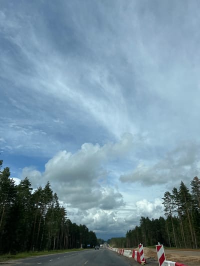 The image captures a view of a road bordered by a dense forest on both sides, likely in the Задорʼеўскі сельскі Савет, Belarus. The road appears to be undergoing construction or repair, as indicated by the presence of temporary barriers and construction materials along the right side. The sky is partially cloudy with dramatic cloud formations and streaks of cirrus clouds. Several vehicles are visible on the road in the distance. The overall scene depicts a rural highway environment with ongoing infrastructure work.