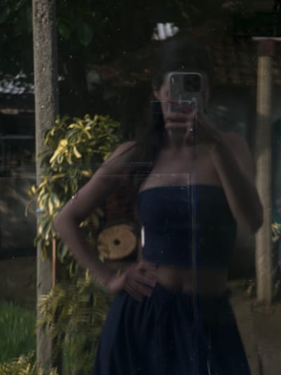 The image displays a reflection on a transparent or semi-transparent surface, likely glass. A person is reflected horizontally across the center of the frame, visible from the torso upwards. They are wearing a dark, strapless top and possibly dark pants or a skirt. Their face is mostly obscured by a smartphone, which they are holding with both hands to capture their reflection. The phone appears to be an iPhone model with multiple camera lenses.

Behind the reflection, an outdoor scene is visible, characterized by lush green and yellow-green foliage from various plants and bushes, suggesting a garden or natural environment. A textured concrete or stone ledge runs vertically along the left and right sides of the frame, serving as a structural element in the background. Near the person's right hand (left in the reflection), a circular, light-colored object, possibly a decorative item or part of a plant pot, is reflected.

The activity taking place is a self-portrait (selfie) or video recording of the person's reflection. The lighting is diffused, indicating daytime, but no specific weather conditions are discernible. The reflective surface shows some minor dust or imperfections. There is no visible text. The setting is an outdoor area in Galabintsi, Bulgaria.