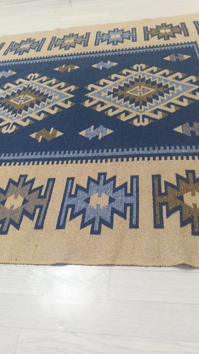 A close-up, top-down, eye-level shot shows a portion of a woven rug with a geometric pattern of blue, tan, and brown colors. The rug is laid out on a light-colored, wood-grain floor. The pattern consists of repeating star-like shapes and zig-zag elements. The rug's edge is slightly frayed. The scene is indoors, and no people are visible. The specific location in the city of Maqat, Kazakhstan, cannot be determined from the image.
