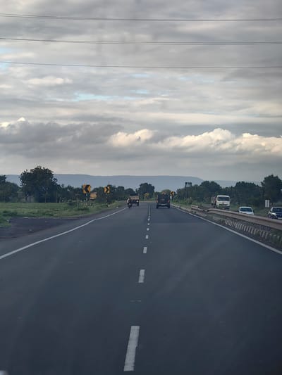 The image depicts a highway in the city of Malkapur, India, under a cloudy sky. The road is asphalt and has white dashed lines marking the lanes. Several vehicles are visible on the road, including a dark SUV, a truck, a white van, a white car, and a motorcycle with a rider. On the side of the road, there are green fields, trees, and hills in the distance. Yellow chevron signs are placed along the roadside, indicating curves in the road. Power lines are strung across the sky.