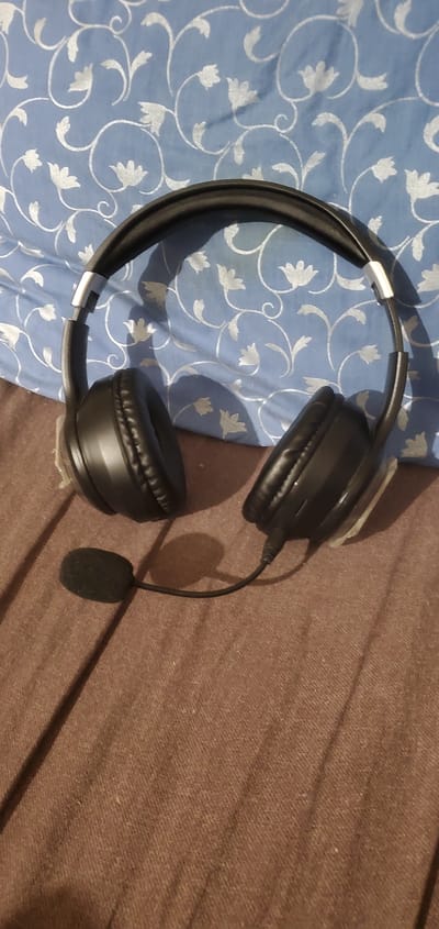 A black headset with a microphone is placed on a brown textured surface, possibly a bedspread or blanket. The headset is a close-up shot, angled slightly to the right. The earcups are padded and black, and the headband is also black with silver adjustable sliders. The microphone is small and covered in black foam, attached to the headset by a flexible wire. In the background, a blue fabric with a white floral pattern is visible, likely a pillow or duvet cover. The lighting casts shadows from the headset onto the surfaces. The image appears to have been taken indoors. This scene is not identifiable as being located in the city of Konakovo, Russia.