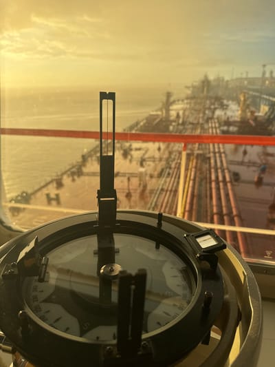 This is a close-up, eye-level shot of a marine compass mounted inside the cabin of a vessel. The compass is brass and black, with a clear glass cover. The needle points towards the South-West, indicated by the "SW" marking on the compass rose. In the background, the vast deck of a large ship stretches out, covered in a network of pipes. The scene is bathed in the warm, golden light of a sunset or sunrise, casting long shadows. The sky is a soft yellow and orange, with a few wispy clouds. The ocean is visible on the left, reflecting the sky's colors. A red railing runs horizontally across the middle of the frame, separating the foreground from the background. The overall impression is one of a serene journey at sea, with the compass guiding the way. There is no indication of Slobodka, Russia, or any people in this image.
