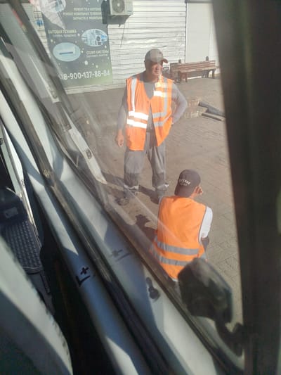In Manhush, Ukraine, two individuals wearing orange high-visibility vests are present outside a building. The person in the foreground, seen from the back, is wearing a dark baseball cap with white lettering and is sitting on the ground. The second individual, facing forward and wearing a baseball cap, stands with one hand on their hip, appearing to look towards the left. In the background, a wooden bench is visible, and a metal rolling shutter is partially open, revealing a white corrugated metal wall. A sign is mounted on the shutter, advertising repair services for televisions and laptops, mobile phones, and split systems, along with a phone number. The scene is illuminated by sunlight, casting shadows on the paved ground.