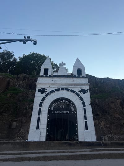 The image shows the Immaculate Conception Grotto of Lourdes in La Esperanza, Honduras. The grotto, built into a rock face, is constructed with white stone, featuring an arched opening enclosed by a black metal gate. A semi-circular inscription, partially visible, reads "INMACULADA CONCEPCION DE LOURDES." Two towers flank the grotto, topped with pointed roofs. A small cross sits atop the structure. Black silhouettes of what appear to be birds are visible on the structure. The surrounding environment includes rock formations, vegetation, and concrete stairs. To the left of the grotto stands a metal pole supporting security camera equipment. A power line is also visible in the sky.