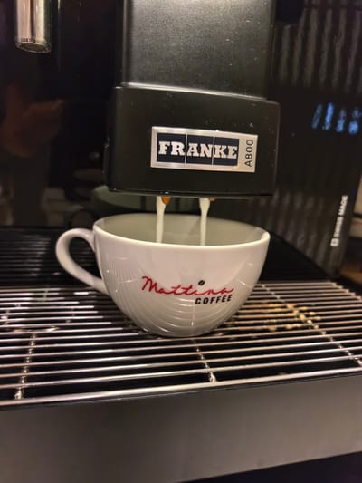 A white "Mattina Coffee" cup is being filled with milk by a black Franke A800 coffee machine on a stainless steel drip tray. The setting appears to be indoors, possibly a cafe or office kitchen. There is no explicit evidence to confirm the location is in Al Matar, United Arab Emirates. No people are visible in the image.