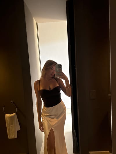 The image shows a young woman in a dark brown corset top and a white silky skirt standing in a bathroom doorway. The bathroom appears to be in a modern apartment or hotel. The woman is holding up a white iPhone to take a selfie. The bathroom is dimly lit, and the woman is illuminated by a bright light behind her. The woman is facing the camera, but her face is obscured by the phone. There is a towel rack with a white towel on it to the left. There is a white switch on the wall to the right of the woman. The coordinates provided (latitude: 31.664, longitude: -7.87) do not appear to correspond to a location in the city of Ouled Jelal, Morocco. However, the location of the image cannot be determined for certain, as it is not possible to discern any landmarks or distinguishing features within the image. The image is a standard selfie style photo, and the focus of the image is the woman. The background is out of focus and only serves to provide a sense of context.