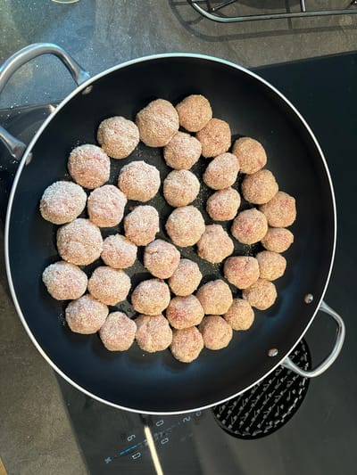 A black, nonstick frying pan sits on an electric stovetop in a kitchen in Celerina/Schlarigna, Switzerland. The pan is filled with 27 evenly spaced, uniformly sized, round, golden-brown, breaded meatball-like items. The pan has a silver handle and is sitting on a black induction cooktop with white control buttons. The stovetop is covered in black laminate with a subtle, repeating geometric pattern.  The camera is facing the pan and the stovetop, from above. The scene appears to be a simple, still life of a pan filled with food in a home kitchen.